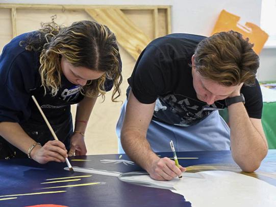 Sonia Lloyd stands at a table, painting a mural alongside another student. 