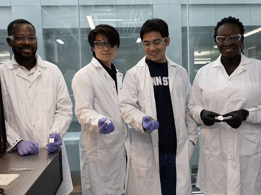 A group of four people wearing lab coats, gloves and goggles pose in a lab while holding parts of a tiny, flat, square-shaped object.