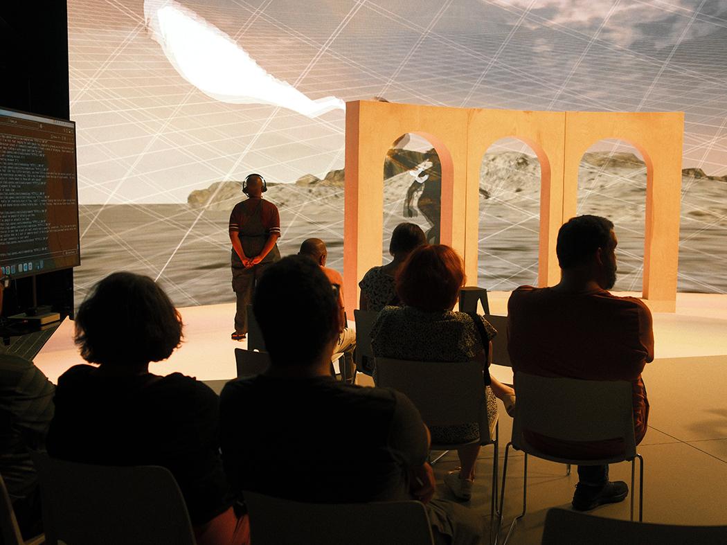 Audience seated in an immersive art installation, watching a person stand before three arch-shaped structures against a projected desert-like landscape with intersecting light lines.