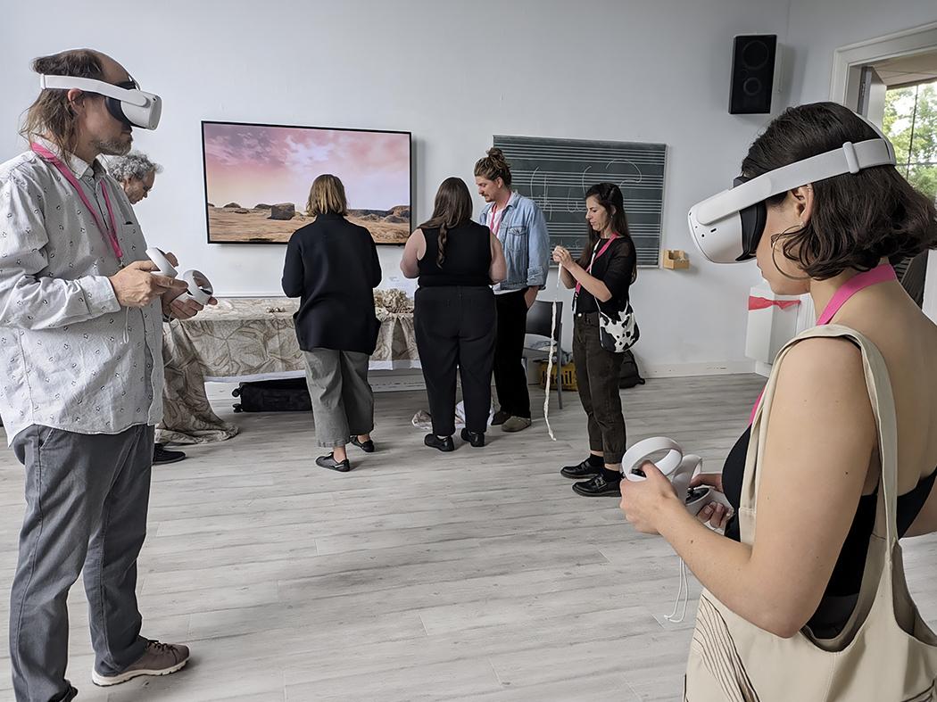 Participants wearing virtual reality headsets and holding controllers in a workshop space, while others gather around a display screen showing a desert landscape.