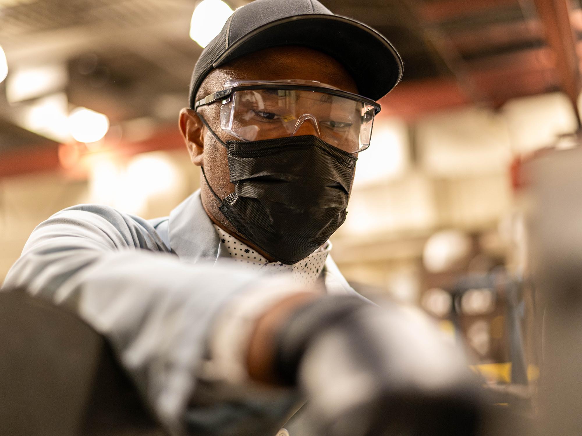A man wearing a black baseball cap, safety goggles and a black surgical mask reaching into the foreground