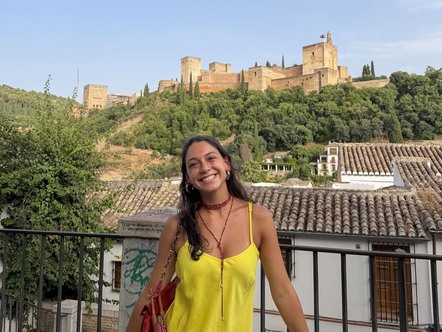 Nicole Schulz, wearing a bright yellow dress, stands in front a railing with the Alhambra in the background.
