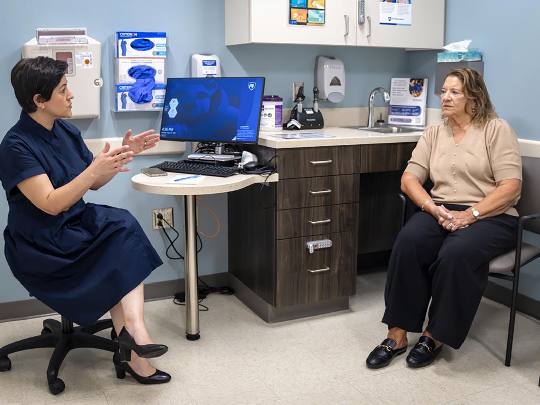 A physician, left, talks to a woman patient in a medical office. The physician gestures with her hands while seated next to a laptop and monitor.  