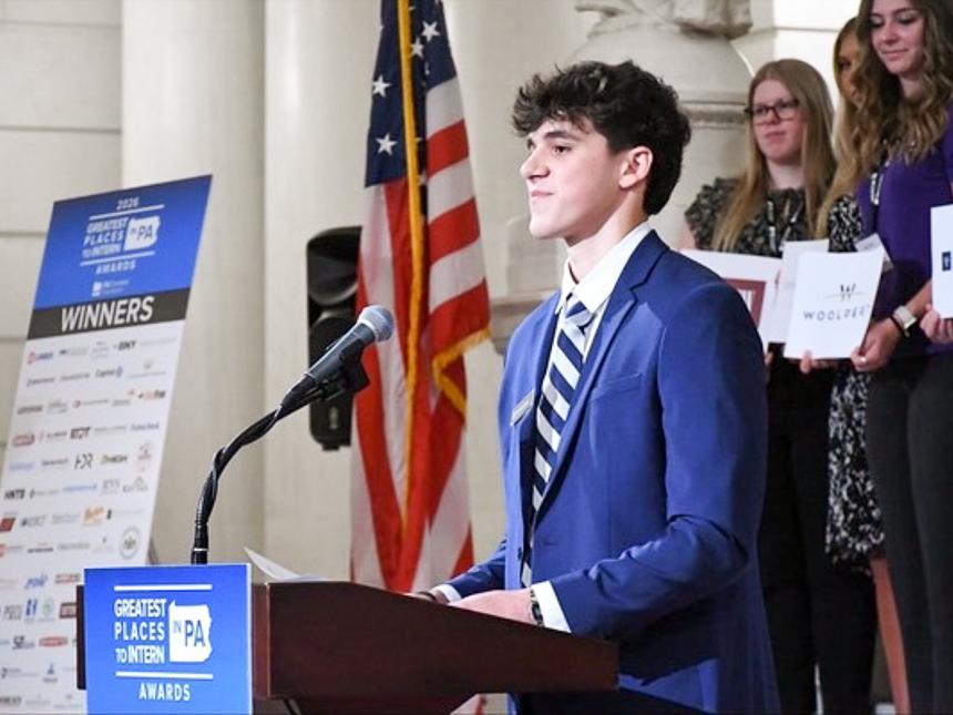 Nikolas Papoutsis in a blue suit delivers a speech at a podium during the Greatest Places to Intern PA Awards event.