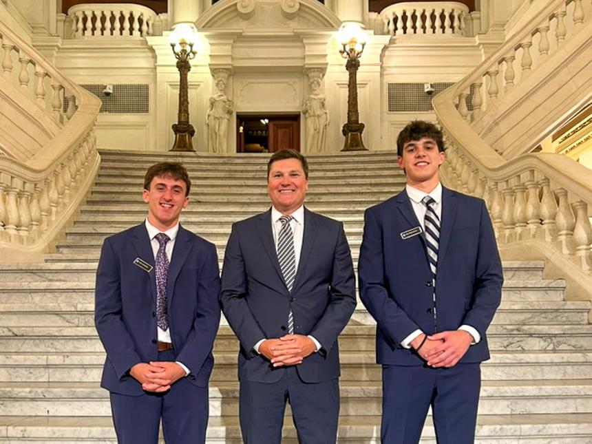 Nikolas Papoutsis, on the right, standing next to a fellow intern and Luke Bernstein, president of the PA Chamber, on steps leading up to a grand entryway of the state Capitol.