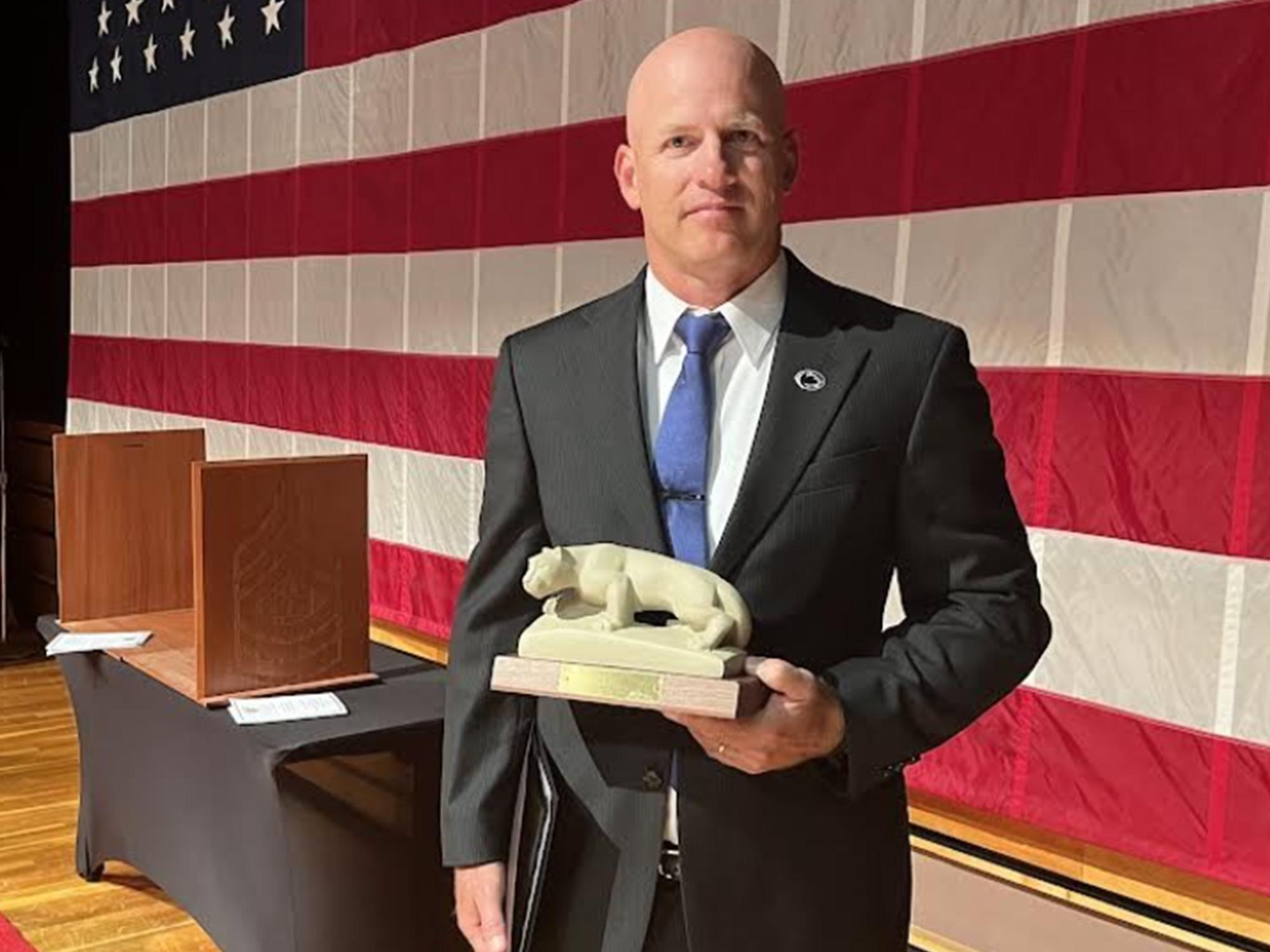 A man wearing a suit poses in front of an American Flag while holding a Nittany Lion minature statue.