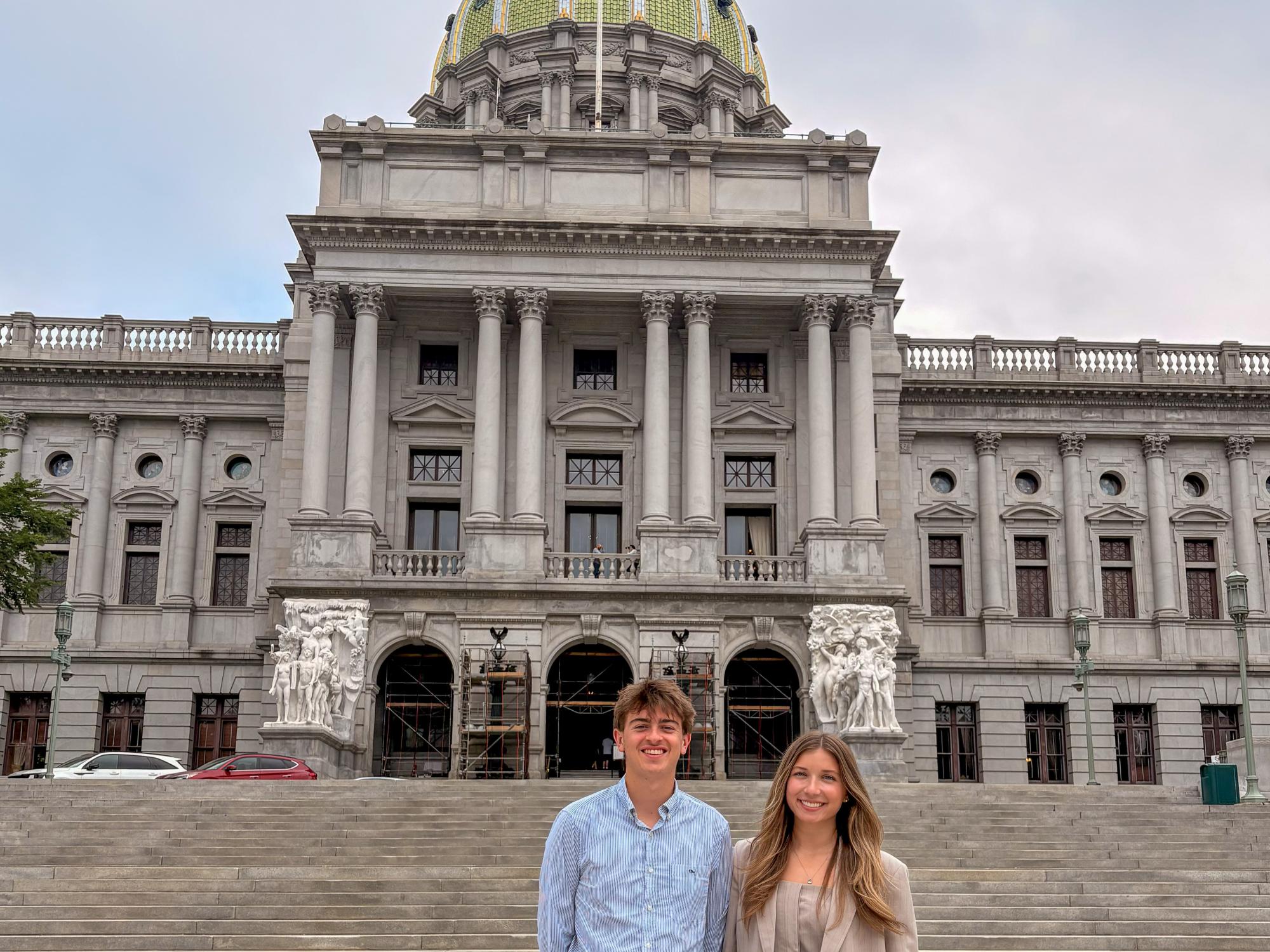 Two students stand in front of a large historic government building with a green dome and flag on top during daytime.