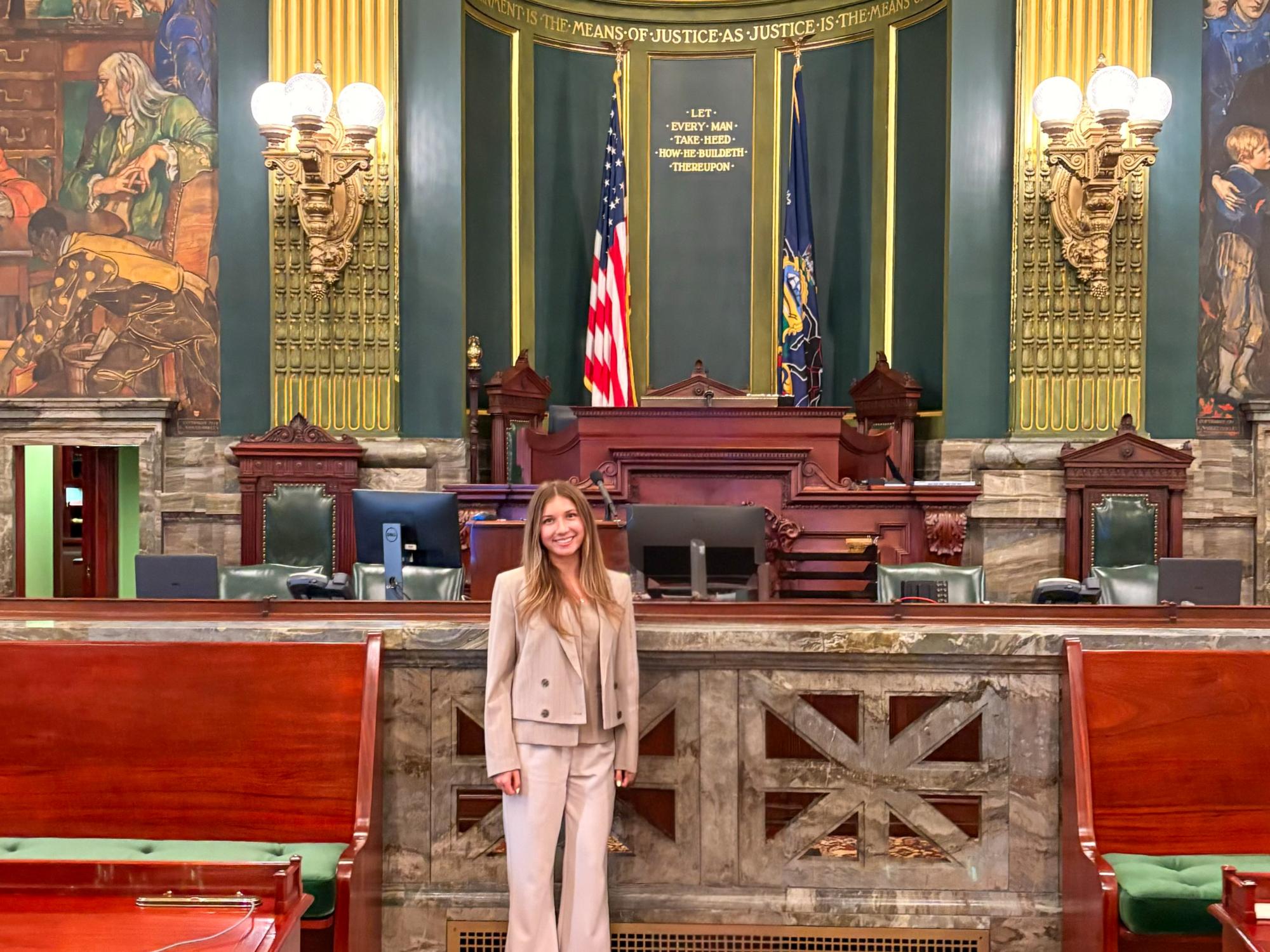 Alexa Pippy, wearing a beige suit, stands on the Pennsylvania Senate floor, with the chamber’s desks and American and Pennsylvanian flags visible behind her. 