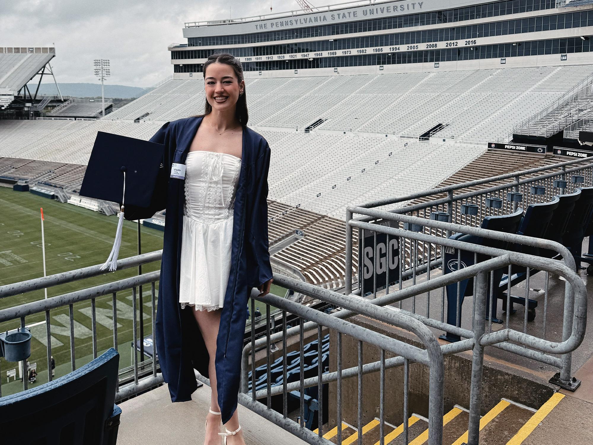 A woman wearing a blue graduation gown poses in front of an empty stadium.