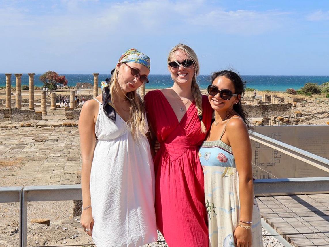 Three people in summer dresses stand in front of ancient stone ruins with tall columns and the sea visible in the background.