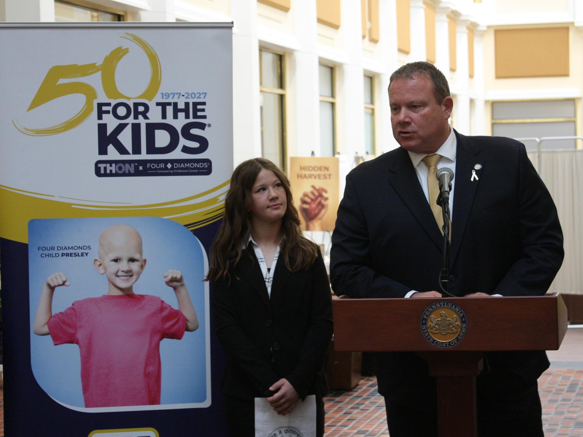 State Sen. Scott Martin speaks at a podium beside a Four Diamonds child and a THON 50 For the Kids standing banner.