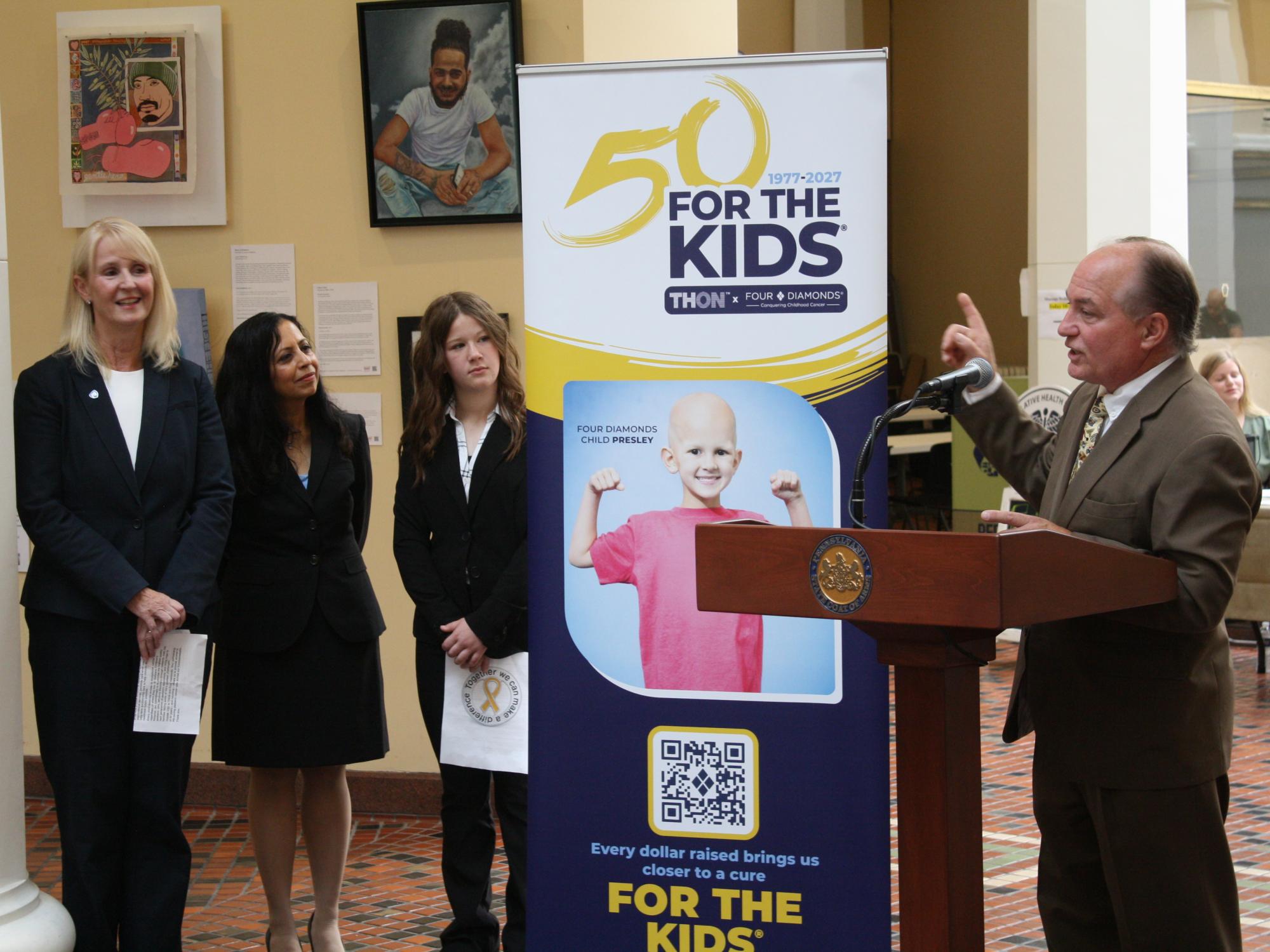 Four people stand beside a podium and a THON and Four Diamonds “50 For the Kids” banner.