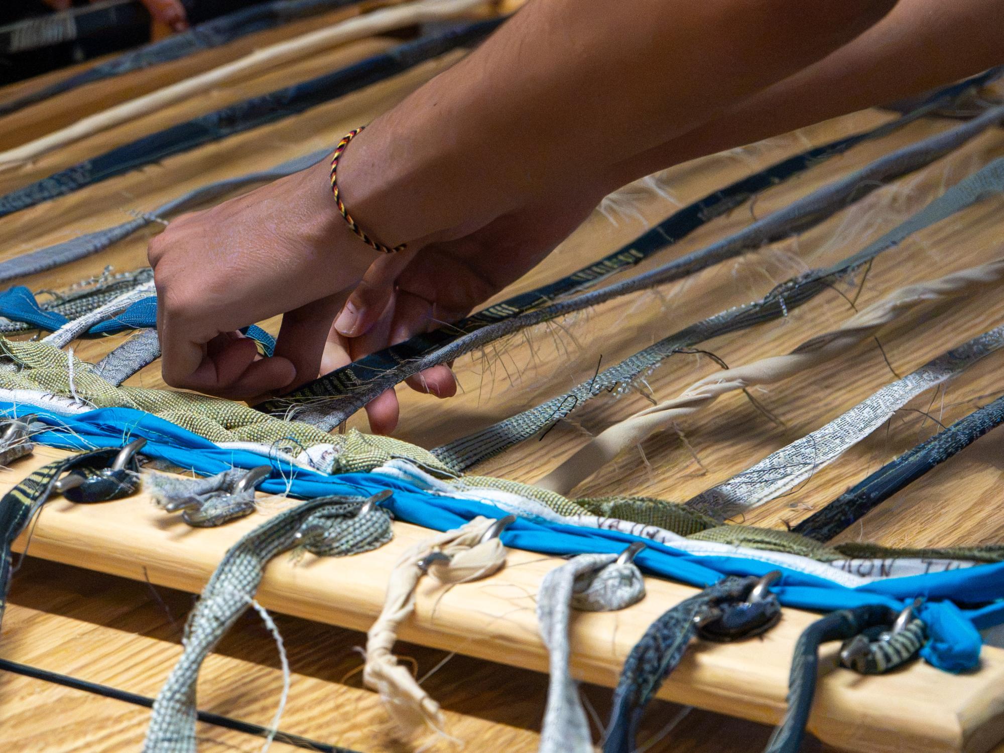 A participant’s hands weave blue, gray and patterned fabric strips through material stretched across a wooden loom.