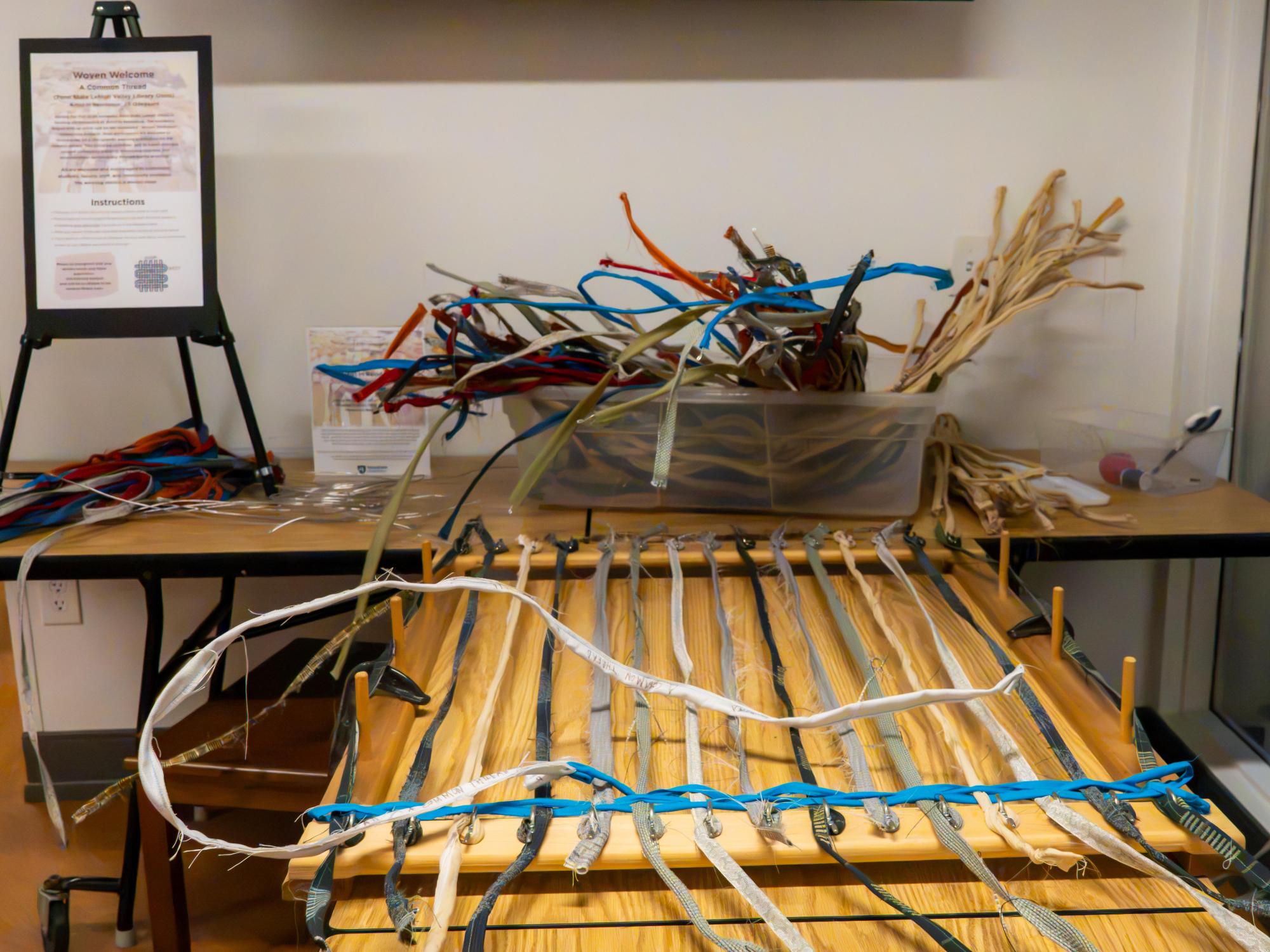 A wooden loom strung with fabric strips sits in front of a container filled with colorful donated materials