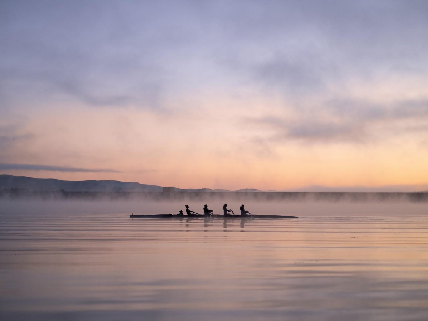 Four individuals row a boat a foggy lake in the shadows at sunrise
