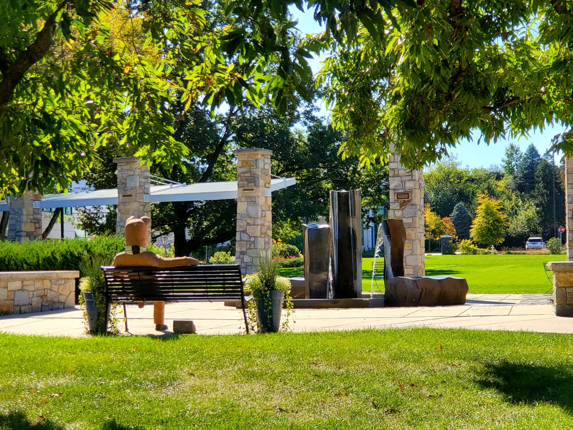 The Nittany Lion sits on a bench looking toward a fountain on a sunny day