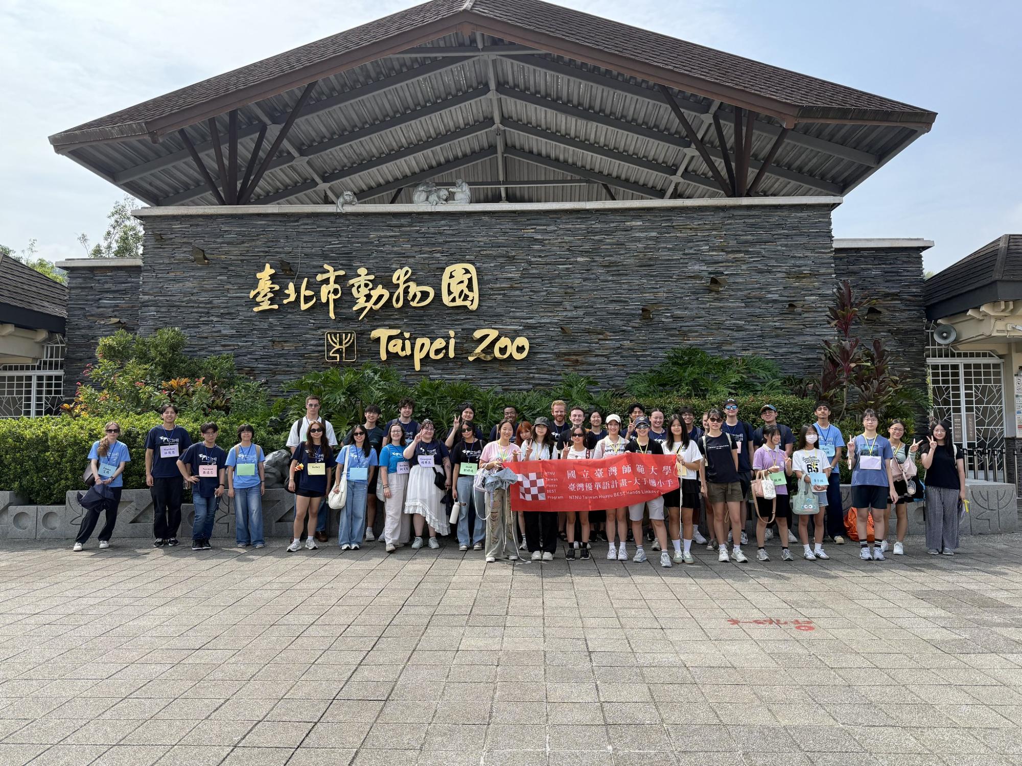 A group stands in front of a building marked "Taipei Zoo" holding a National Taiwan Normal University Huayu Best Program banner