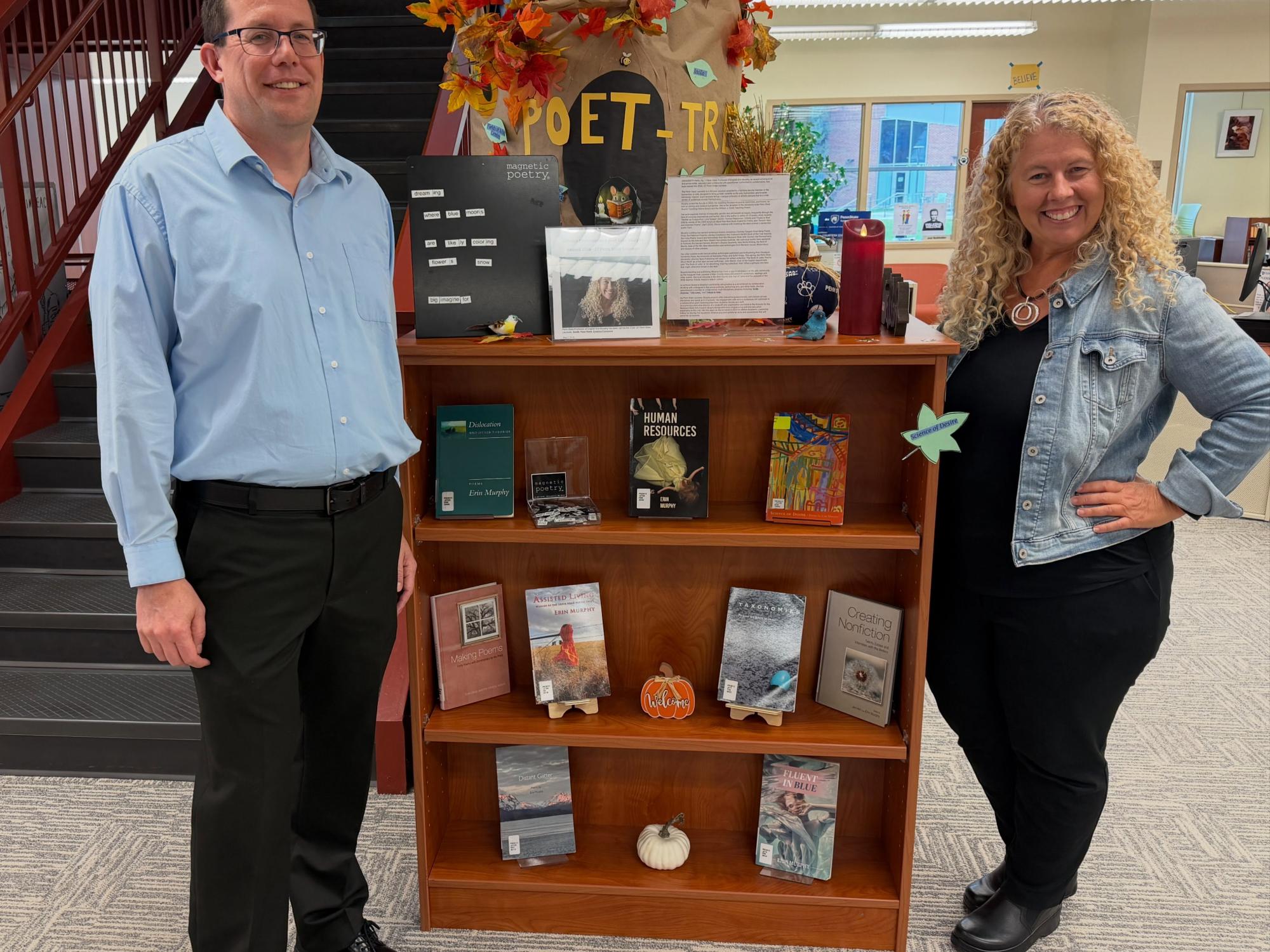 Tom Reinsfelder and Erin Murphy smile while standing on opposite sides of a bookshelf displaying Murphy’s books, poetry activities and fall decorations.