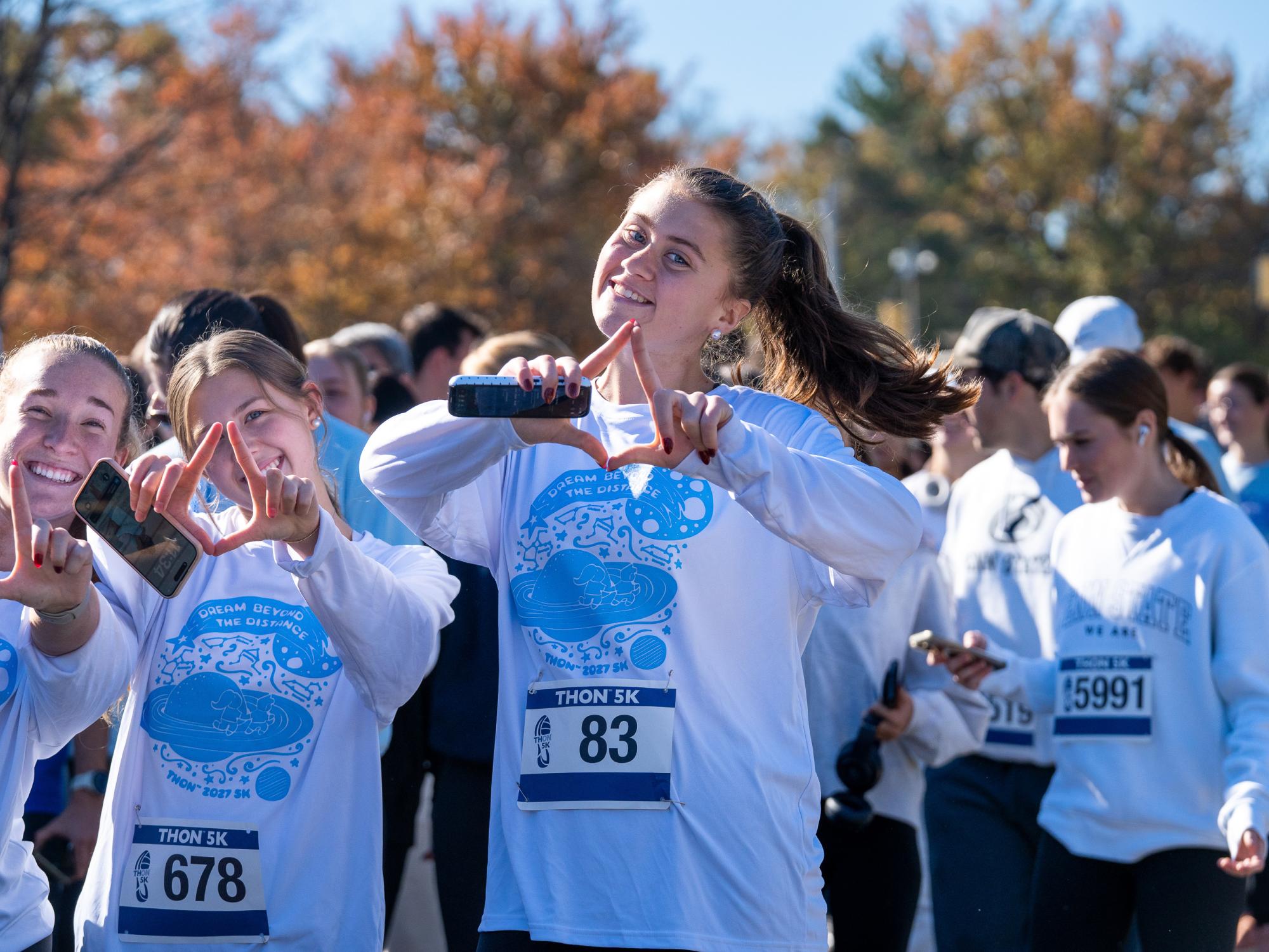 Three smiling participants wearing white THON 5K shirts and race bibs form diamond shapes with their hands while holding phones. A crowd of runners and walkers gathers behind them on Penn State's University Park campus, with colorful autumn trees in the background.