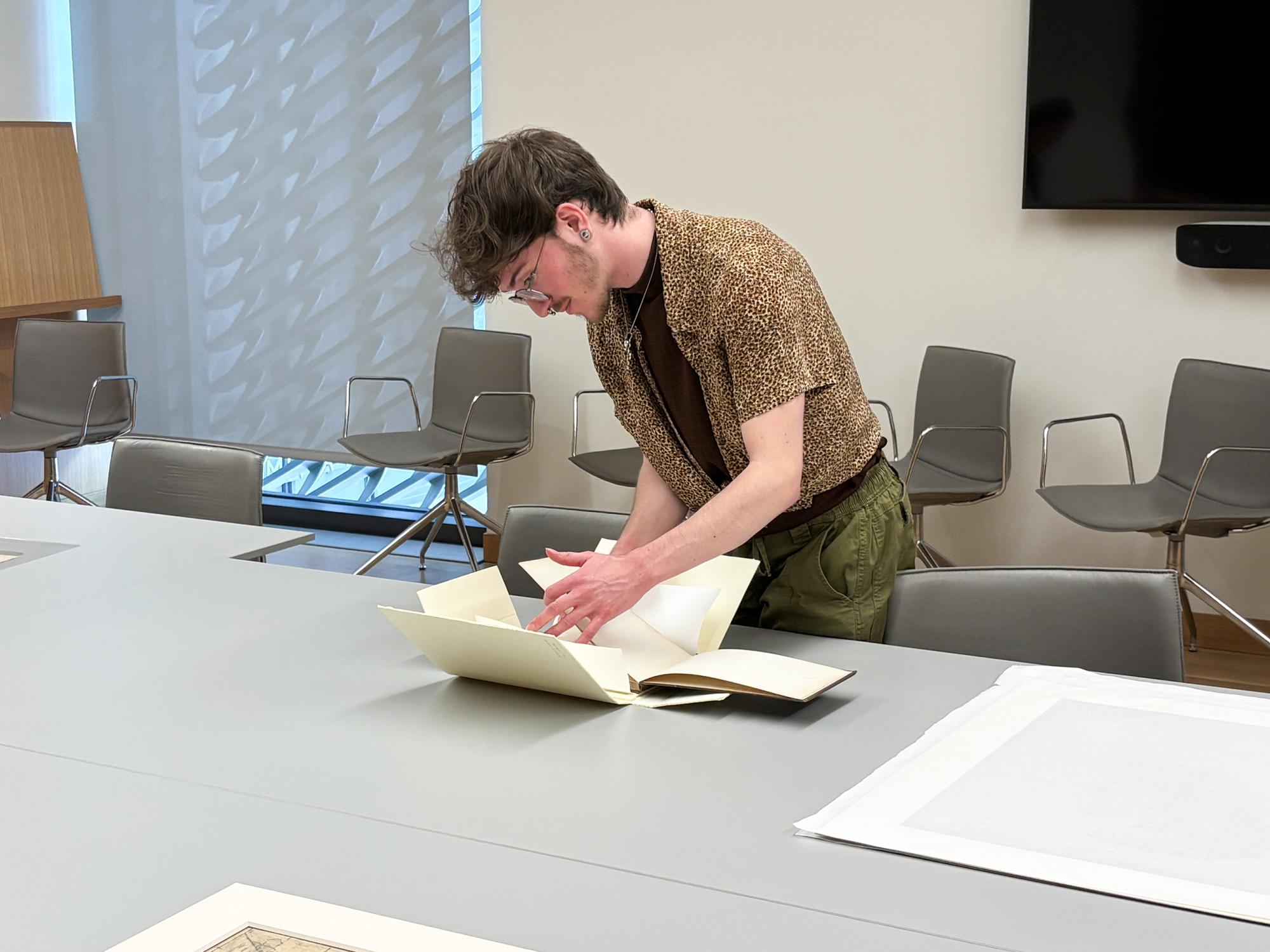 A student leafs through an old sketchbook that sits on a big gray table. Other artworks like across the table near him.