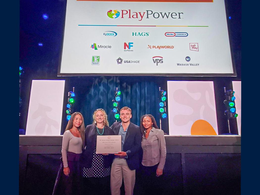 Four people stand on a stage beneath a large PlayPower sponsor sign and colorful stage lights. Two people hold a framed certificate while the group poses for a photo. Various company logos appear on the sign above them.