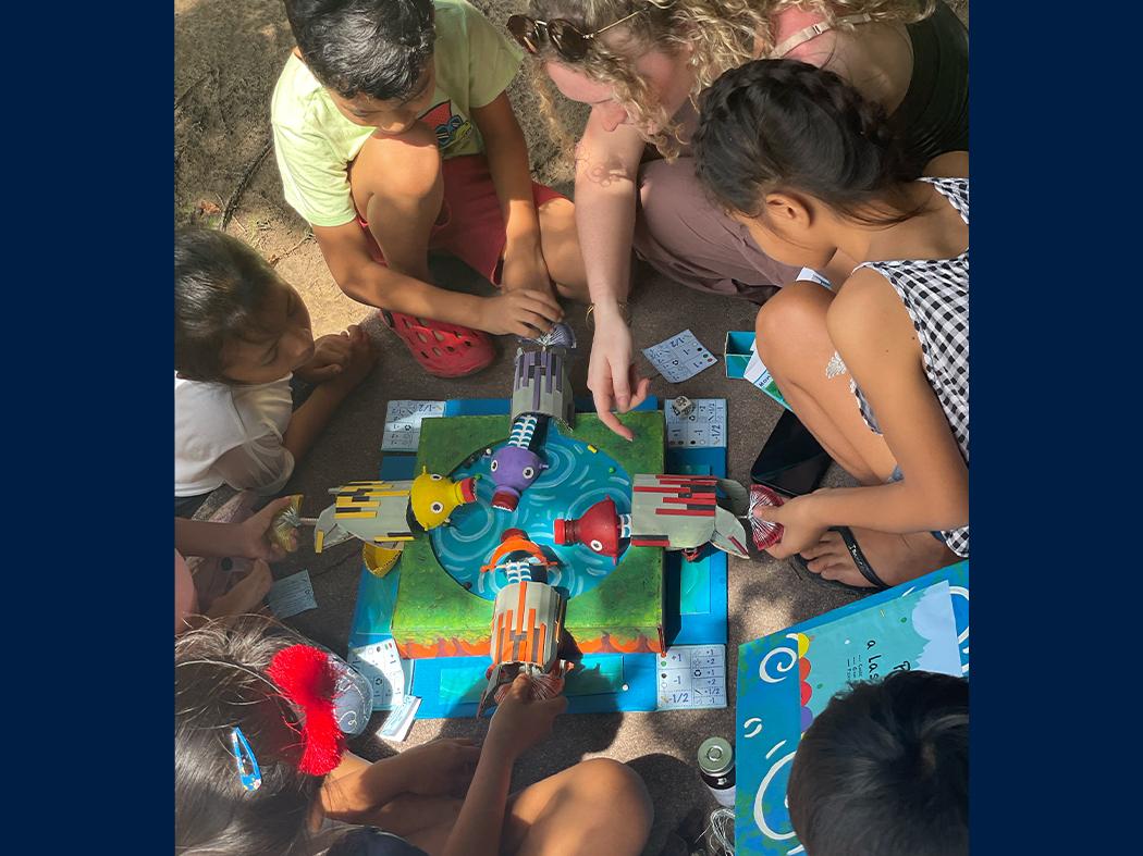 Several children and adults sit around a colorful tabletop game on the ground. They hold or adjust game pieces around a circular blue-and-green board, with cards and a children’s book scattered nearby.