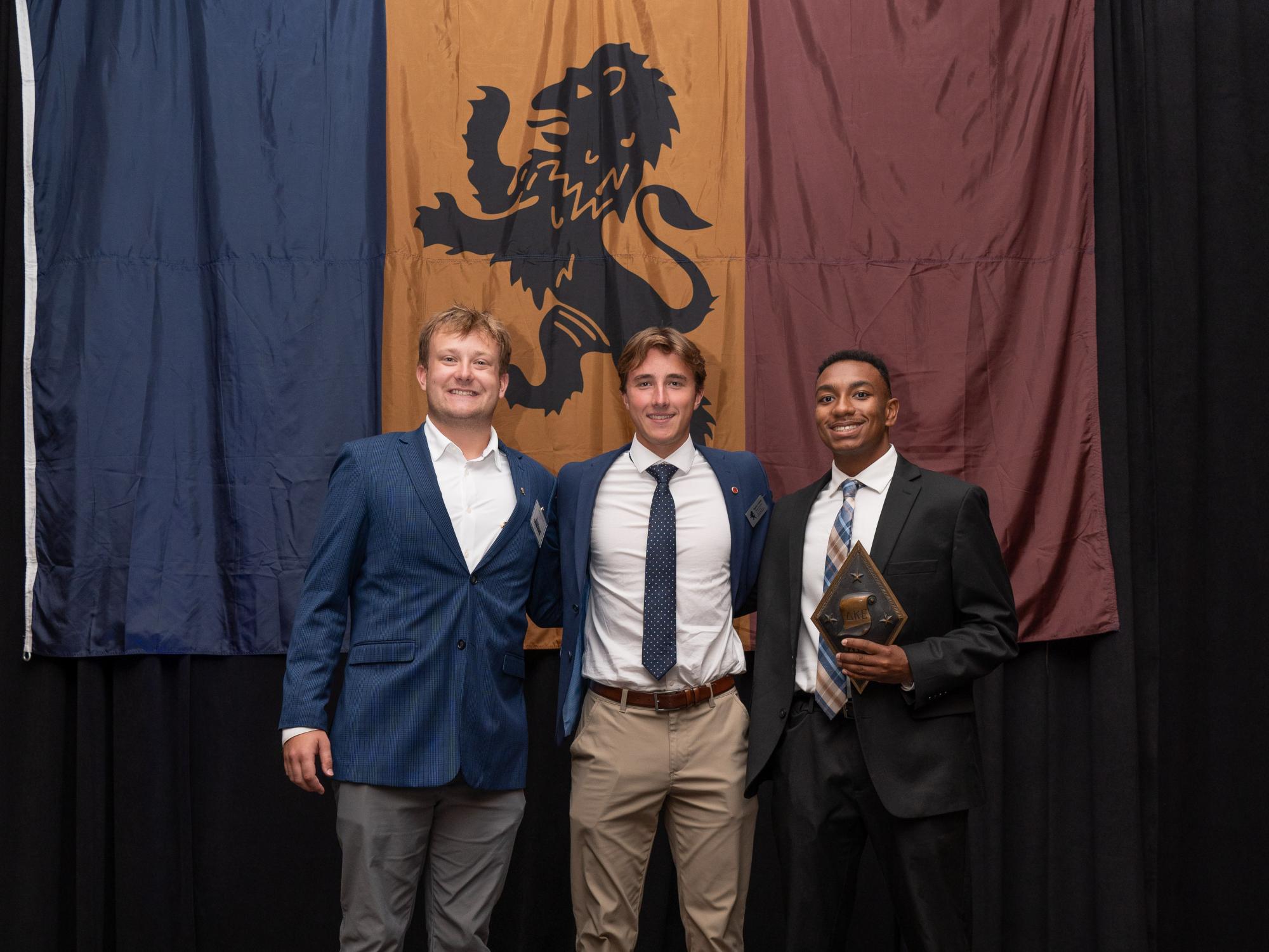 Three people in suits stand on a stage in front of a blue, gold, and maroon banner with a lion emblem. Jaylen Davis holds a plaque.