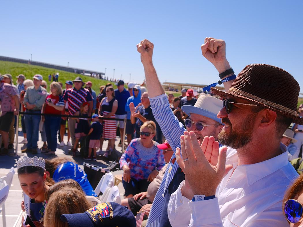 A man in a woven hat and sunglasses claps in the foreground as spectators cheer behind him, including a person raising both fists.