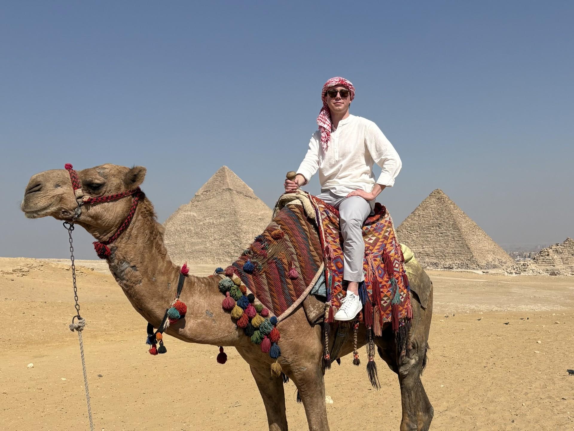 A man sitting on a camel. The pyramids sit in the background.