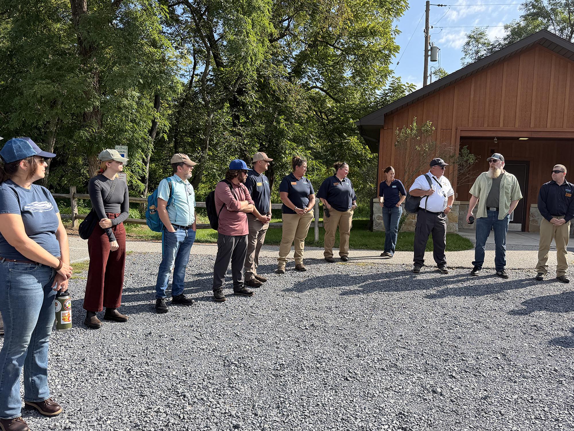 Eleven people in outdoor gear, many in sunglasses, standing as part of a large circle in an outdoor setting