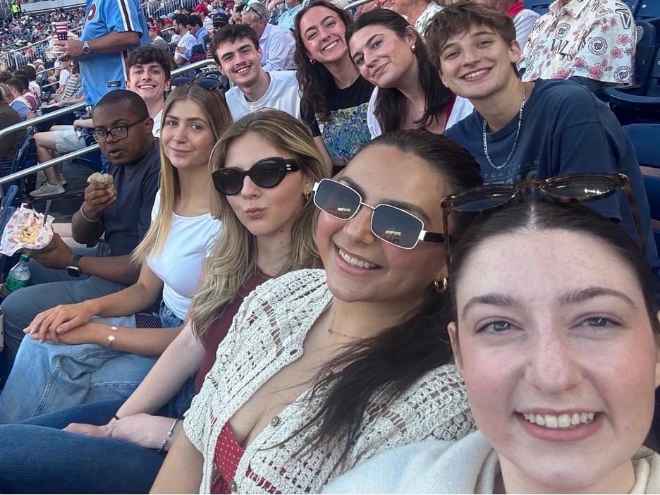 Group of ten students smiling and enjoying a sunny day at a crowded outdoor baseball stadium.