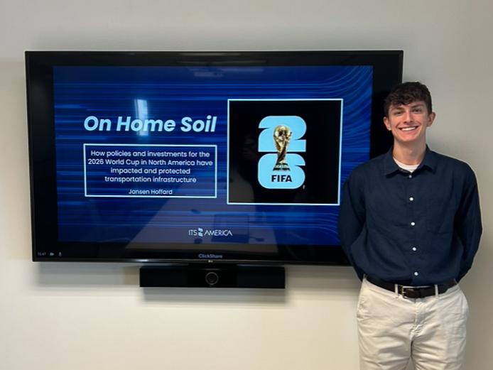 Jansen Hoffard stands beside a large indoor screen display showing a presentation titled “On Home Soil”.