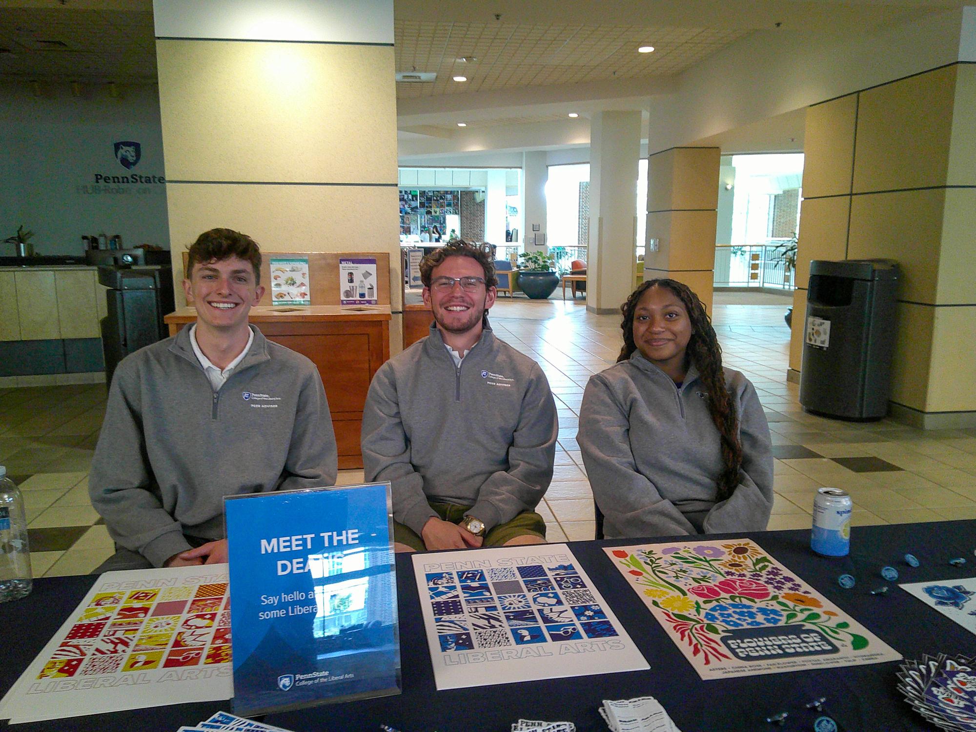 Three individuals wearing matching gray Penn State Liberal Arts sweaters sit behind a table with colorful signs and posters.