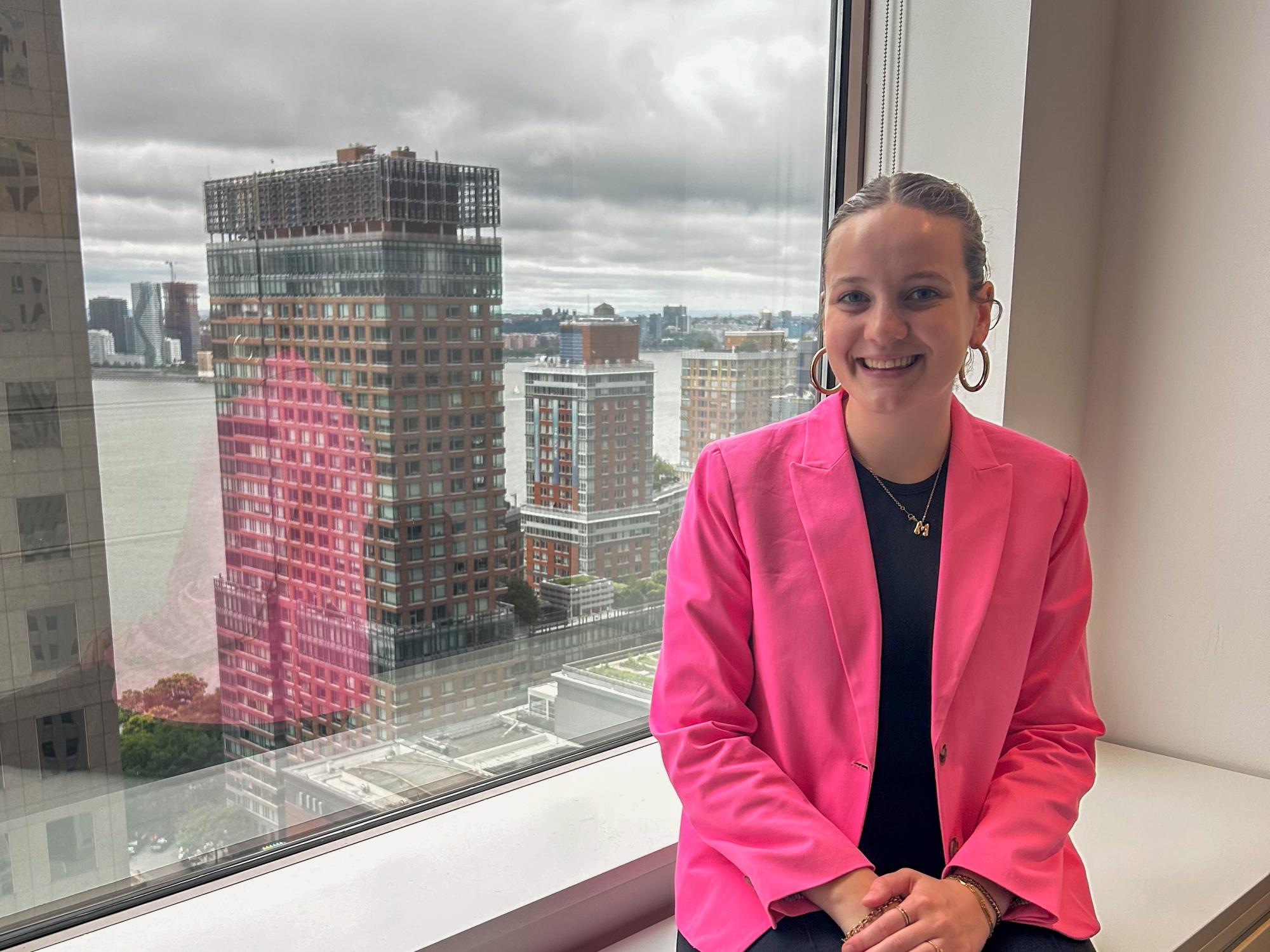 Madeline Shanafelt in a bright pink blazer sitting on a windowsill with a cityscape and river visible outside the window on a cloudy day.