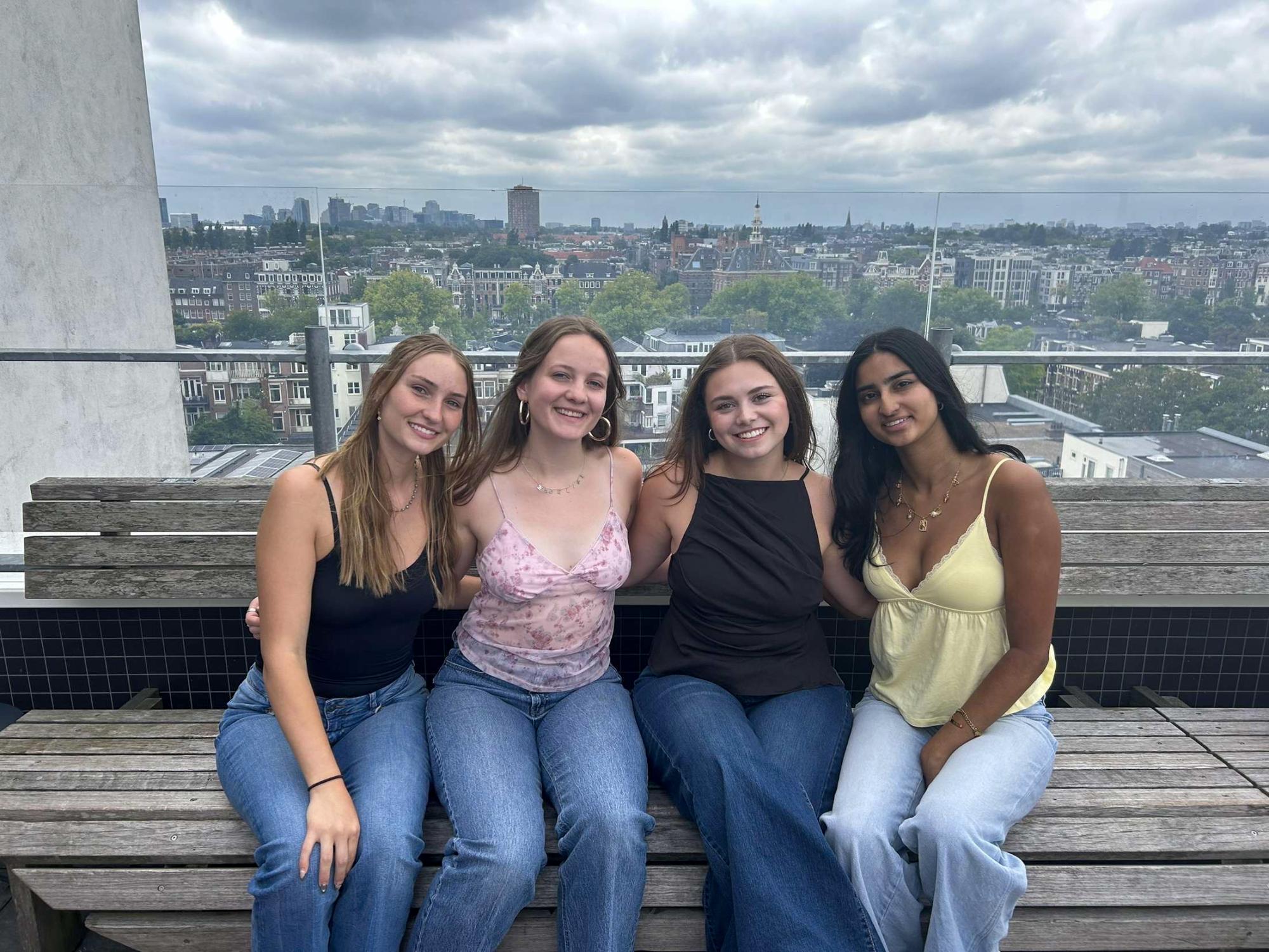 Four young women, including Madeline Shanafelt, sitting closely on a rooftop bench with a cityscape under cloudy skies behind them.