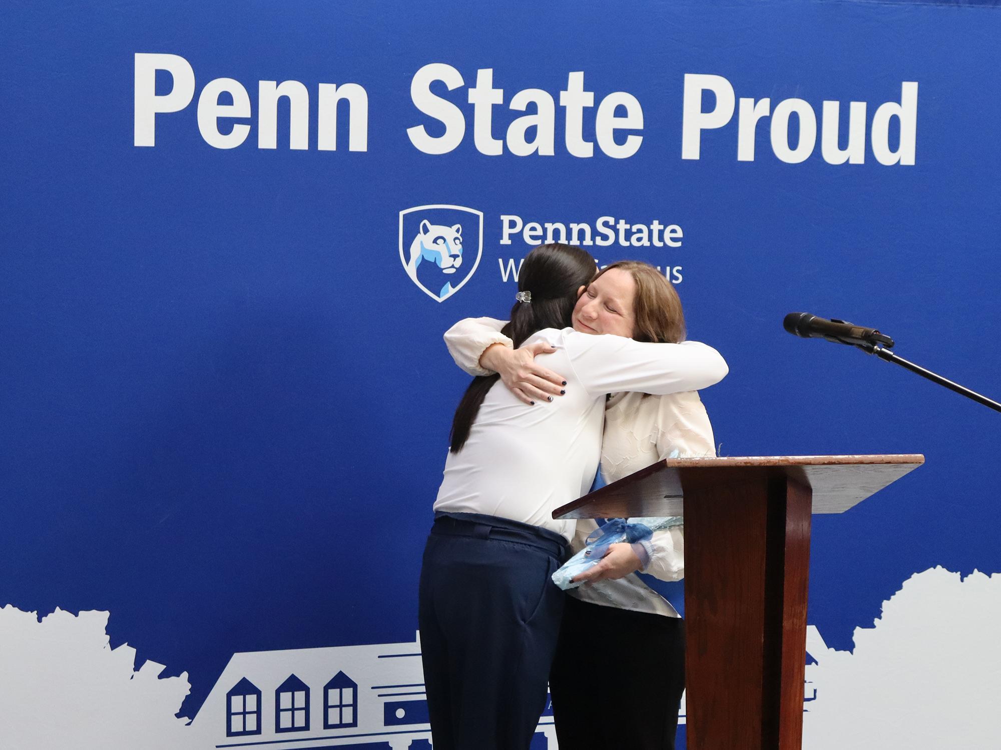 Two women hug in front of a blue Penn State photo background.