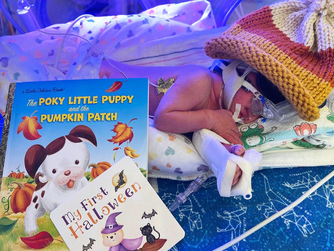 Maximus Coelho lies in an incubator in the NICU wearing a knit cap and with a brace on his arm. Two children’s books are propped up next to him: “The Poky Little Puppy and the Pumpkin Patch” and “My First Halloween.”