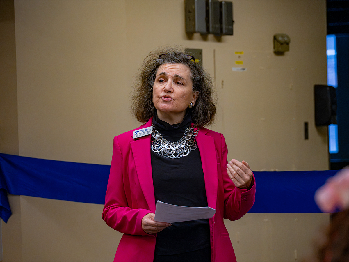 Rosemary Martinelli, wearing a bright pink blazer, holds notes and gestures while speaking in front of a blue ribbon.