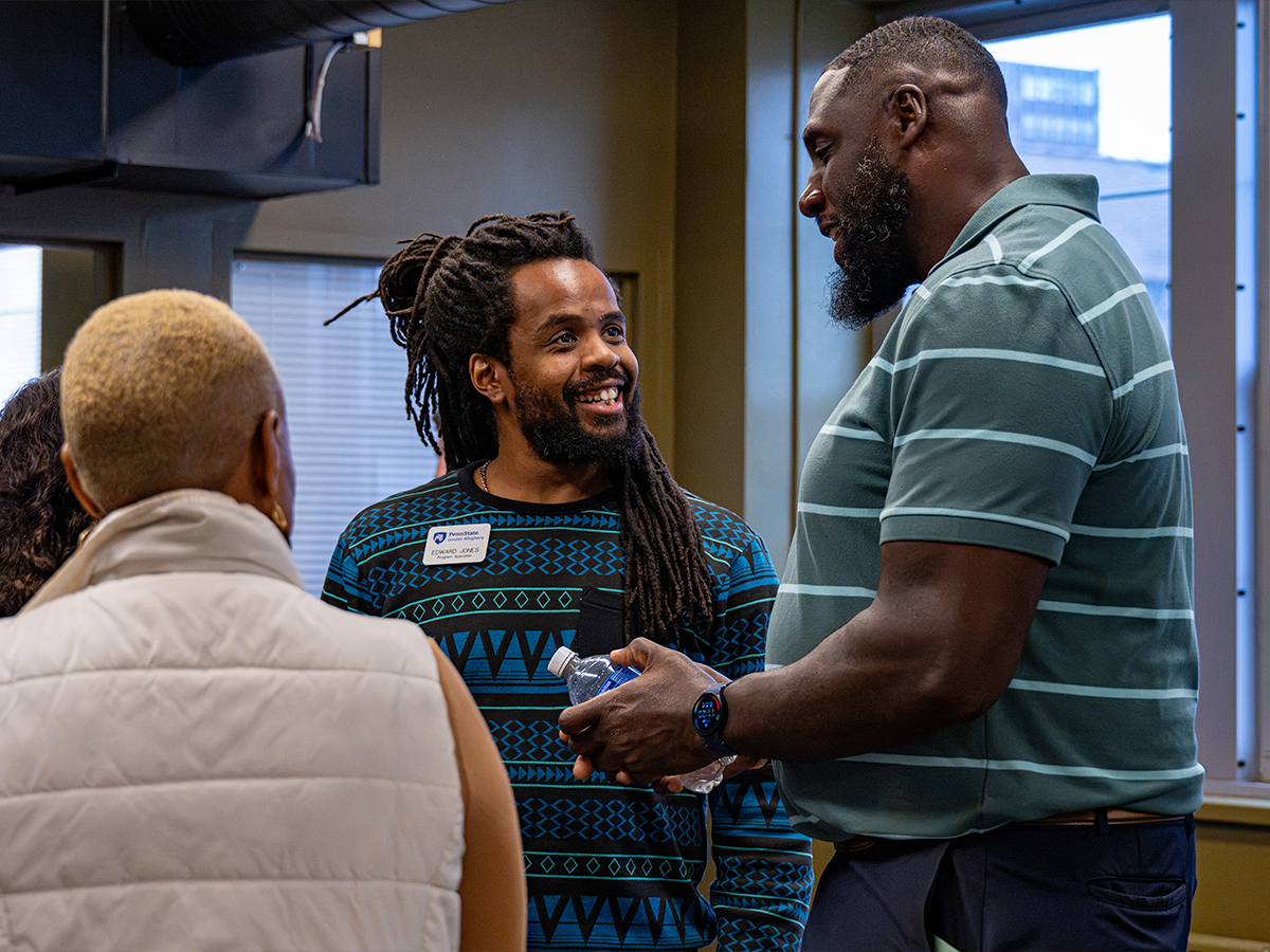 Attendees converse indoors, with a smiling man in a blue patterned shirt facing a man in a green striped polo and a person in a light-colored vest.