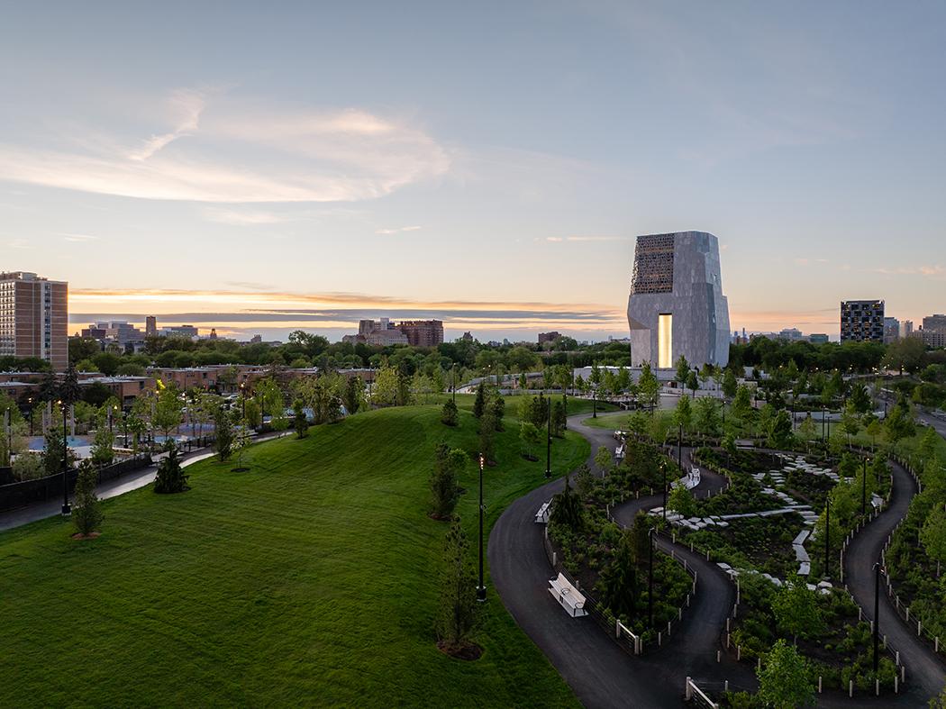Aerial view at sunset of a modern tower with a tall illuminated opening, set amid landscaped grounds, winding roads, and trees, with a city skyline in the distance.