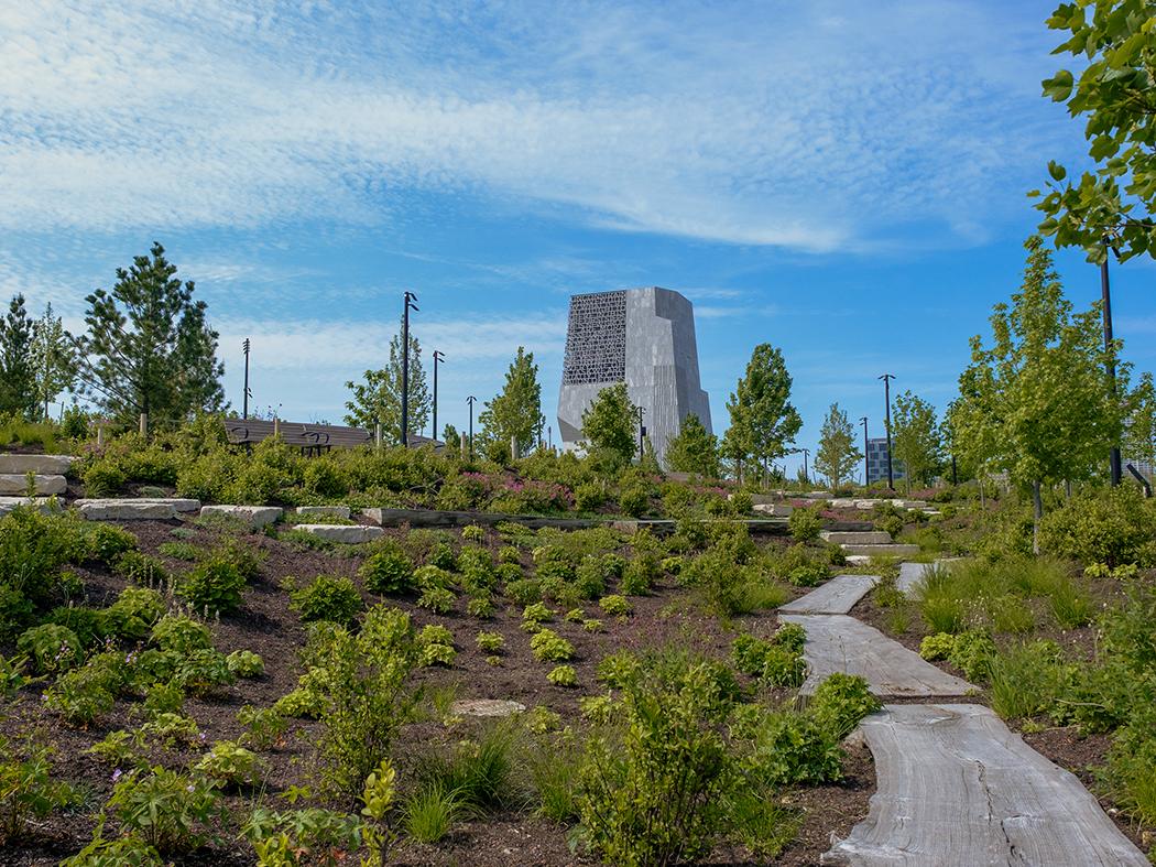 A stone path winds through low native plants toward a modern gray tower on a landscaped rise beneath a bright blue sky.