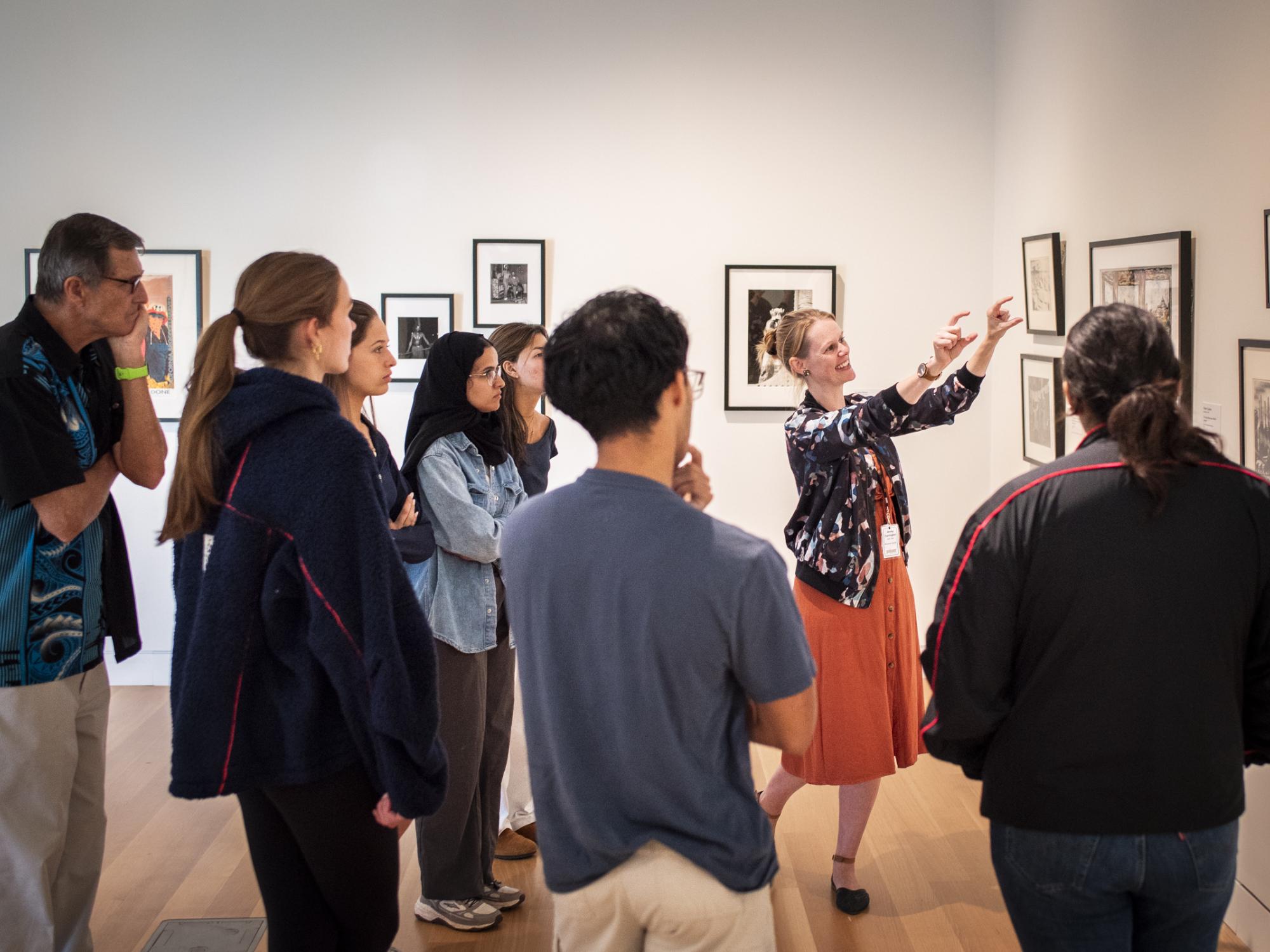 A diverse group of students on a guided tour in a museum. The tour guide has her arms outstretched and is pointing to the artwork she is discussing.