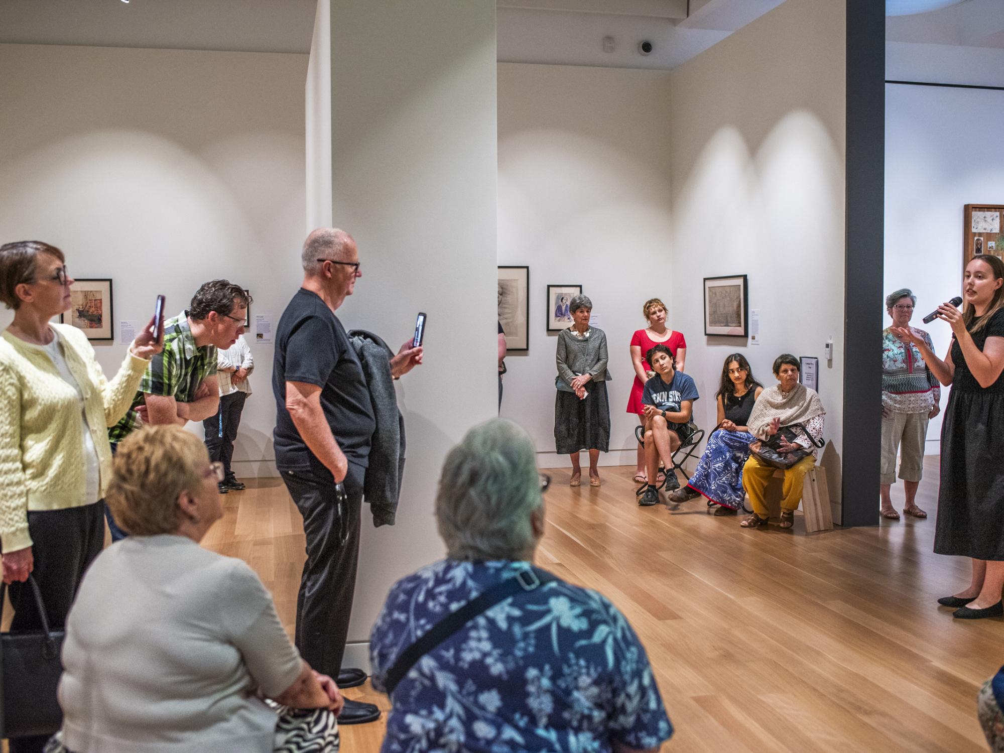A student gives a talk in an art gallery to a large group of people. Some sit on stools and many stand.