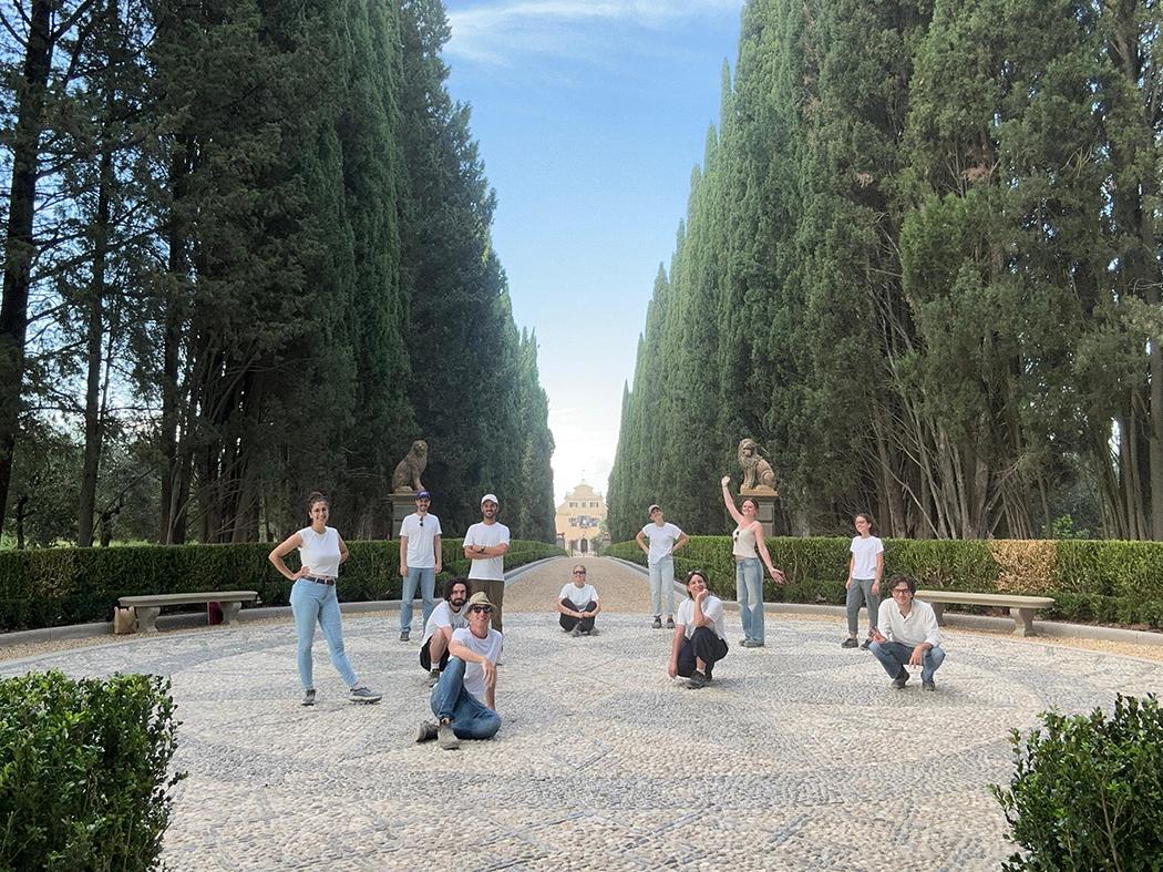 Twelve people pose in a formal courtyard lined with tall cypress trees, clipped hedges, stone benches, and statues, with a historic building visible in the distance.