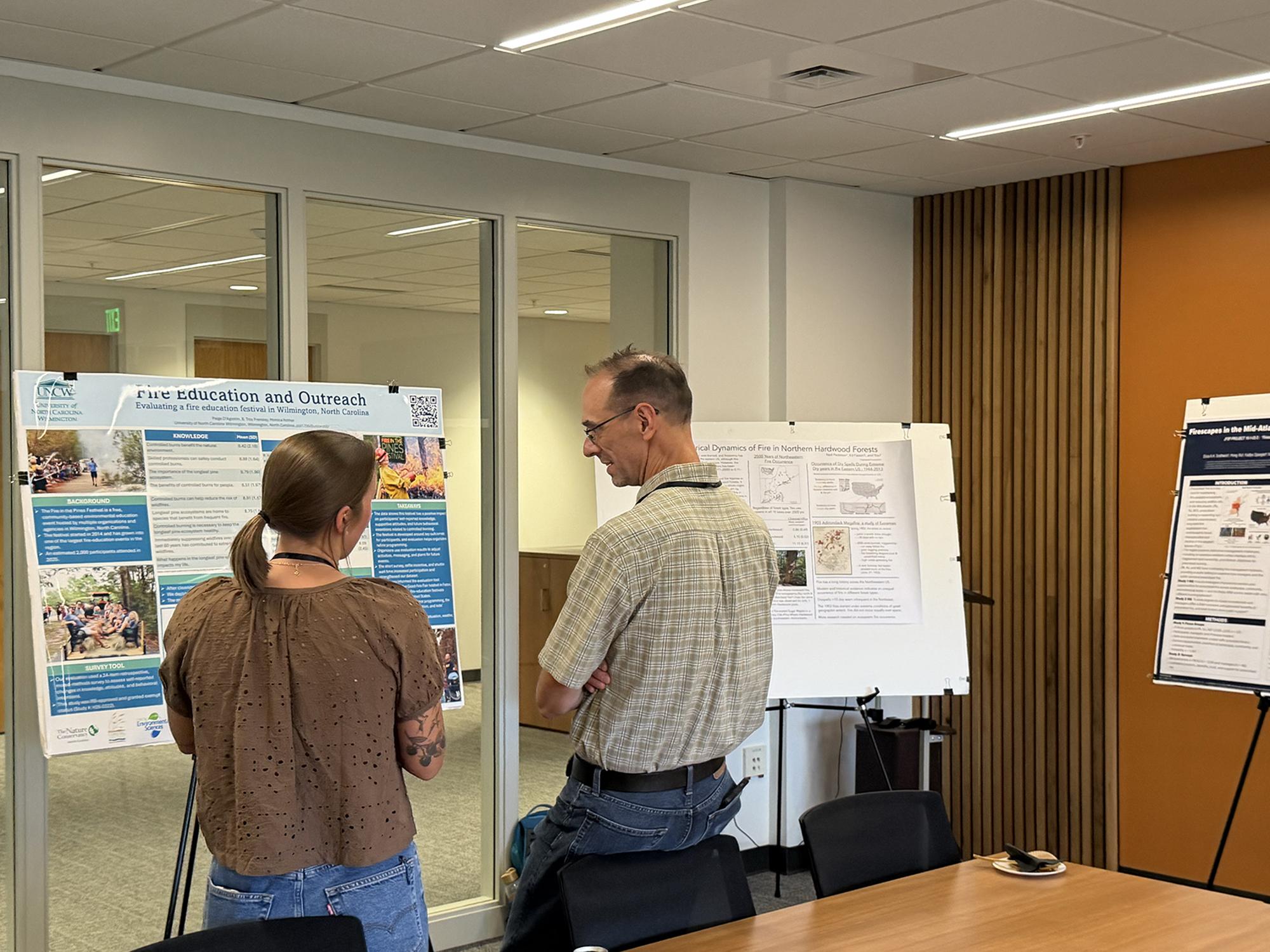 Two people talking in front of an academic poster with blue text boxes. Two other posters in the background