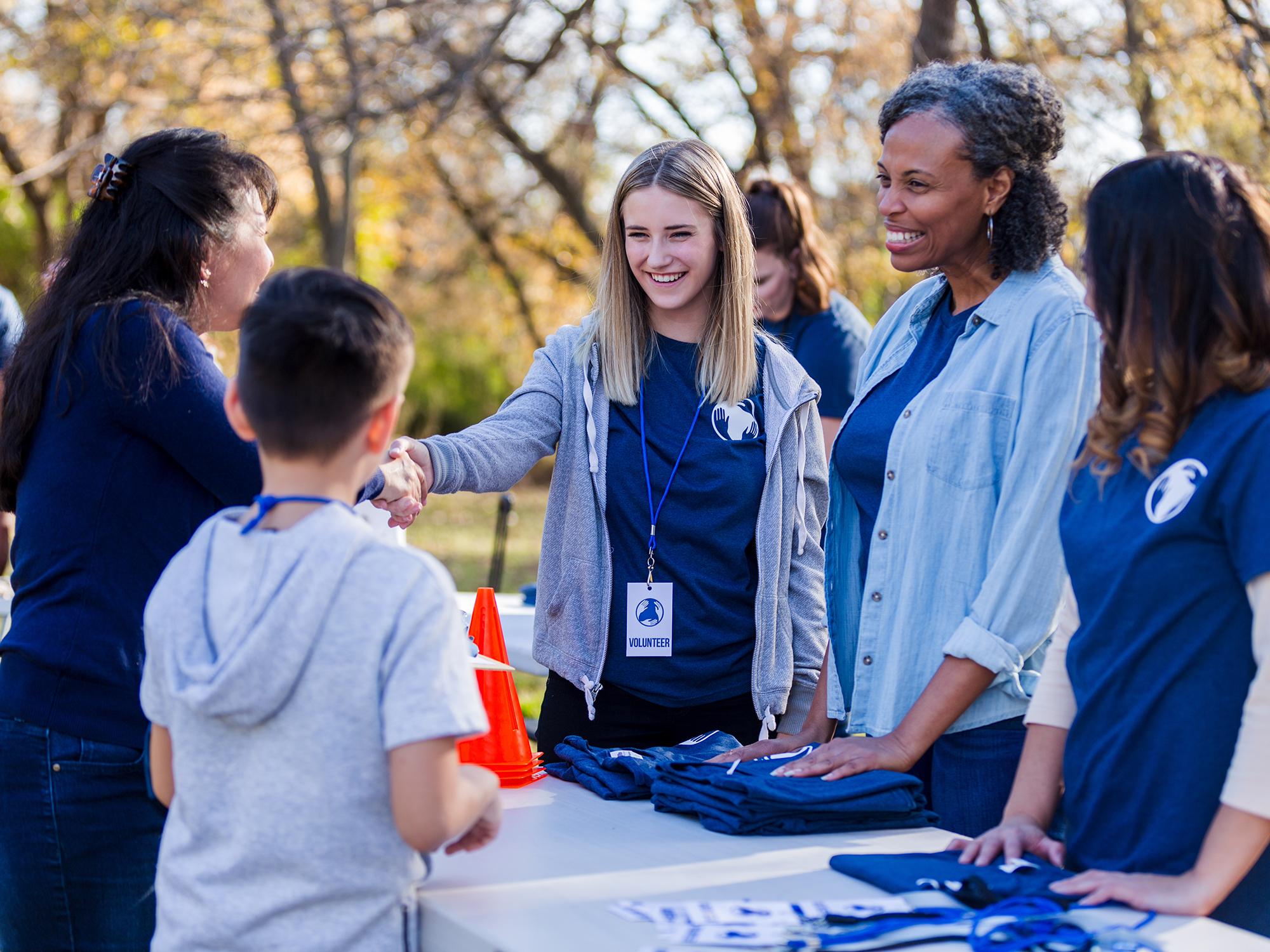 Volunteers at an outdoor event greet and interact with a child and other participants, distributing materials.