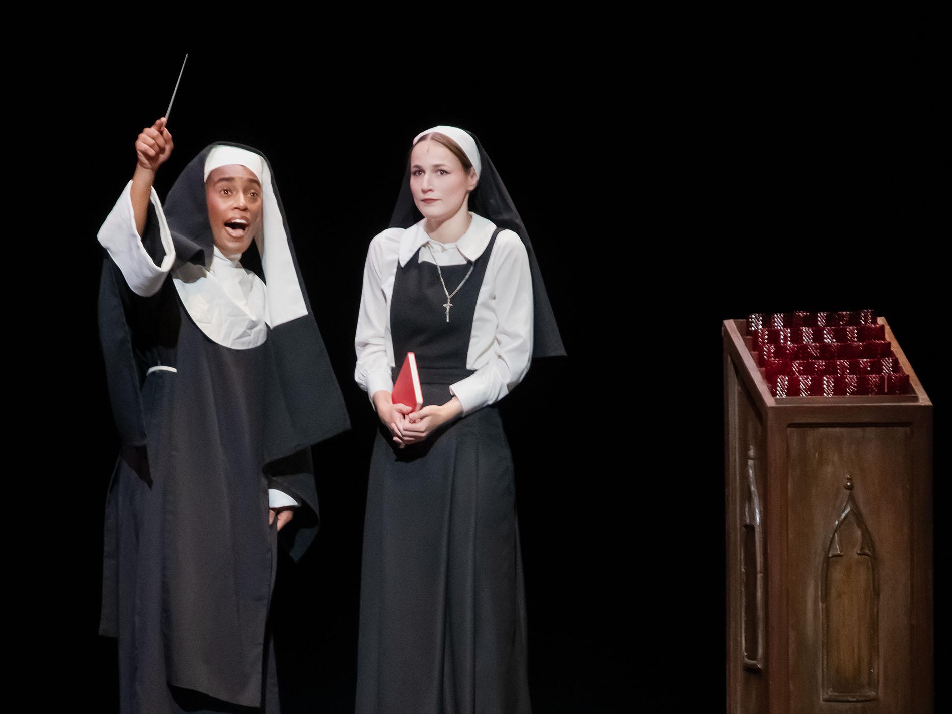 Two performers dressed as nuns perform on stage next to a podium