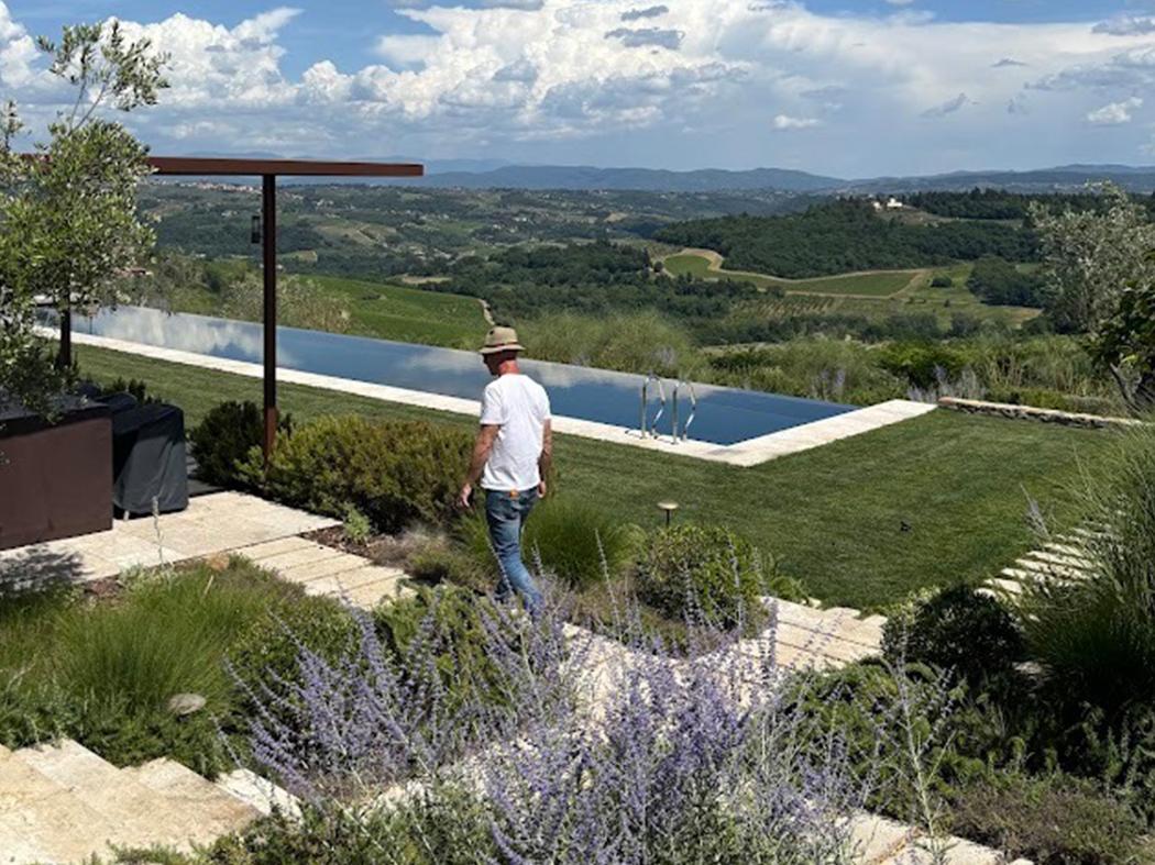 A man walks through a landscaped garden beside a long reflecting pool, with lavender, ornamental grasses, and rolling green hills stretching toward distant mountains under a partly cloudy sky.