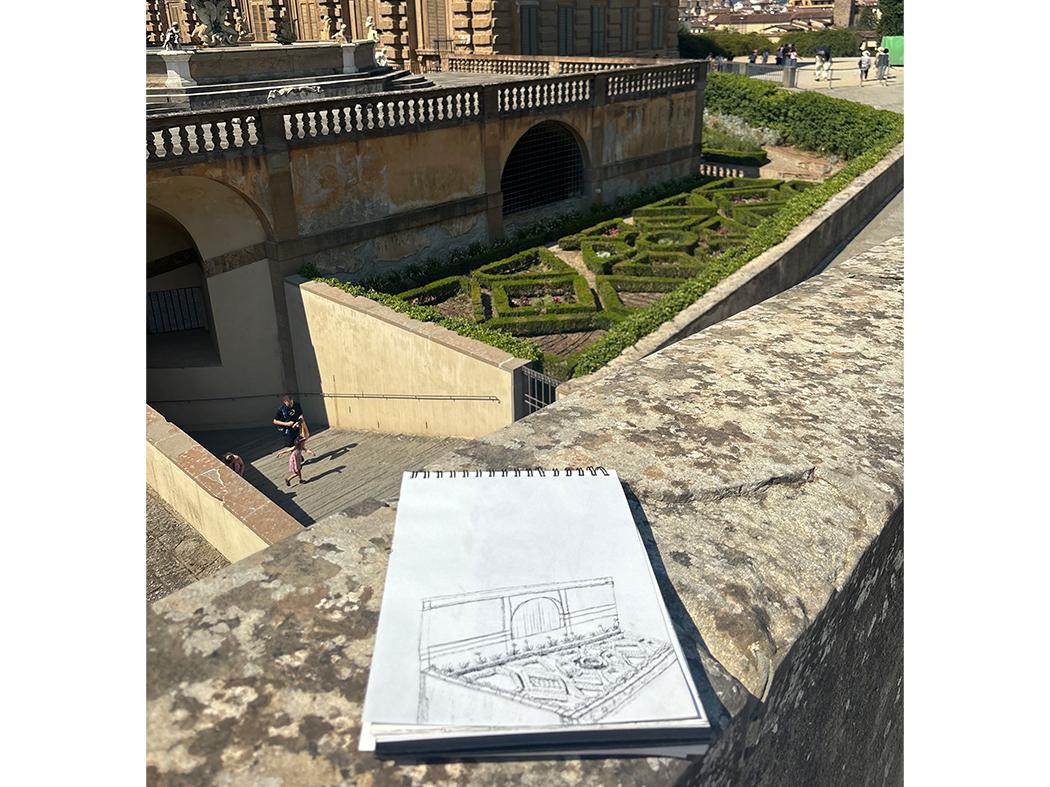 Sketchbook with a pencil drawing of a formal geometric garden and arched historic building, resting on a stone ledge overlooking the actual garden and courtyard below.