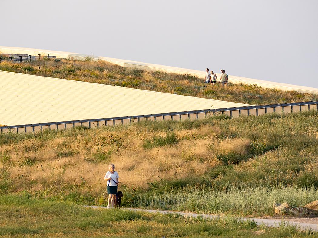 A person walks along a path through tall prairie grasses below a sweeping, grass-covered roof; three people stroll along the upper path.